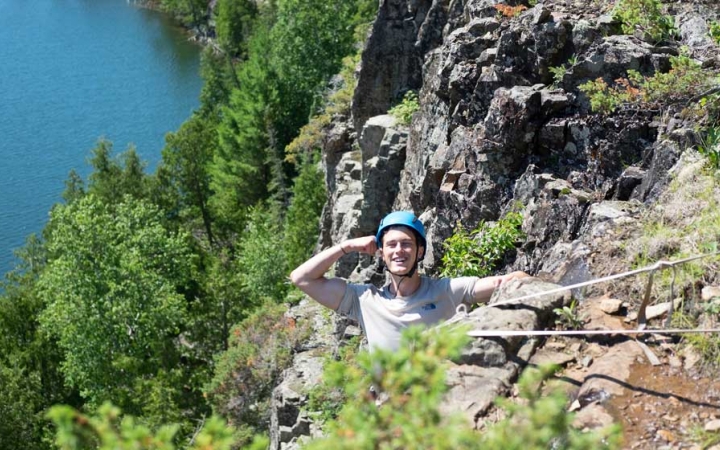 A person wearing safety gear is secured by ropes as they salute the camera while rock climbing, high above a body of water. 